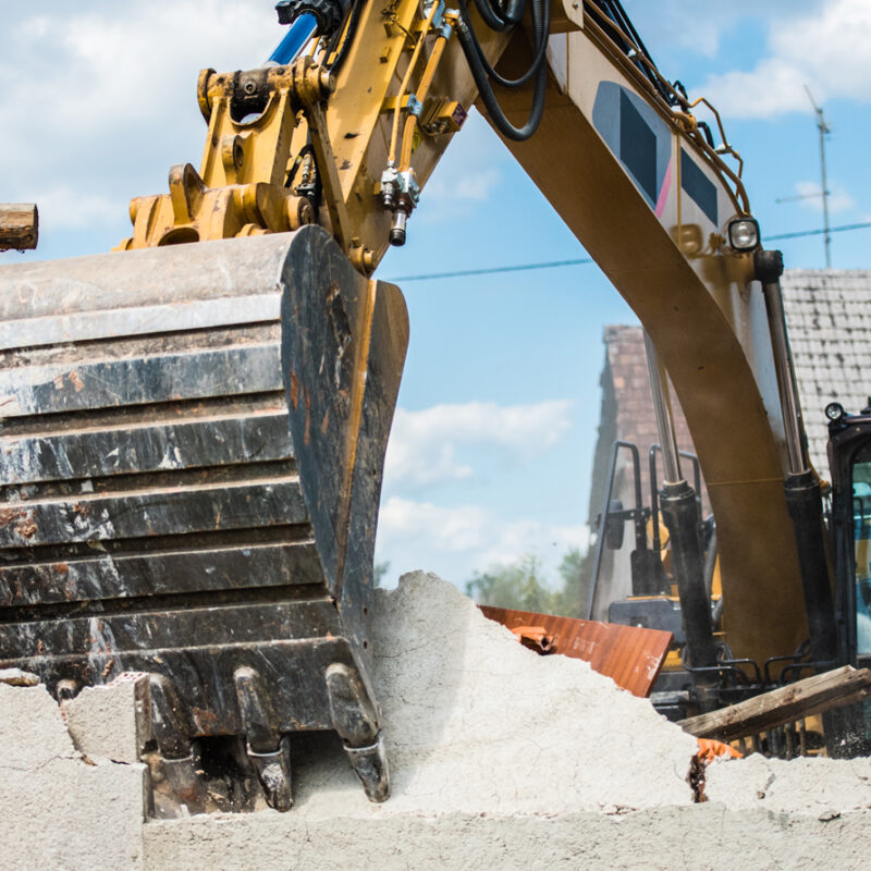 Heavy construction vehicle tearing down the wall of a house with the scoop in the foreground