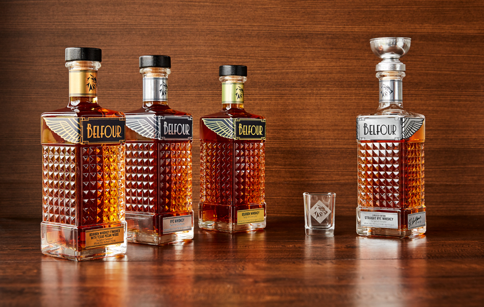 Belfour Pecan Whiskey with metallic copper label, Rye Whiskey with metallic silver label, Bourbon with metallic gold label and limited edition Whiskey with metallic silver label and silver hockey trophy topper on a polished wood table with a polished wood background and a shot glass featuring the diamond eagle logo mark of Belfour Spirits