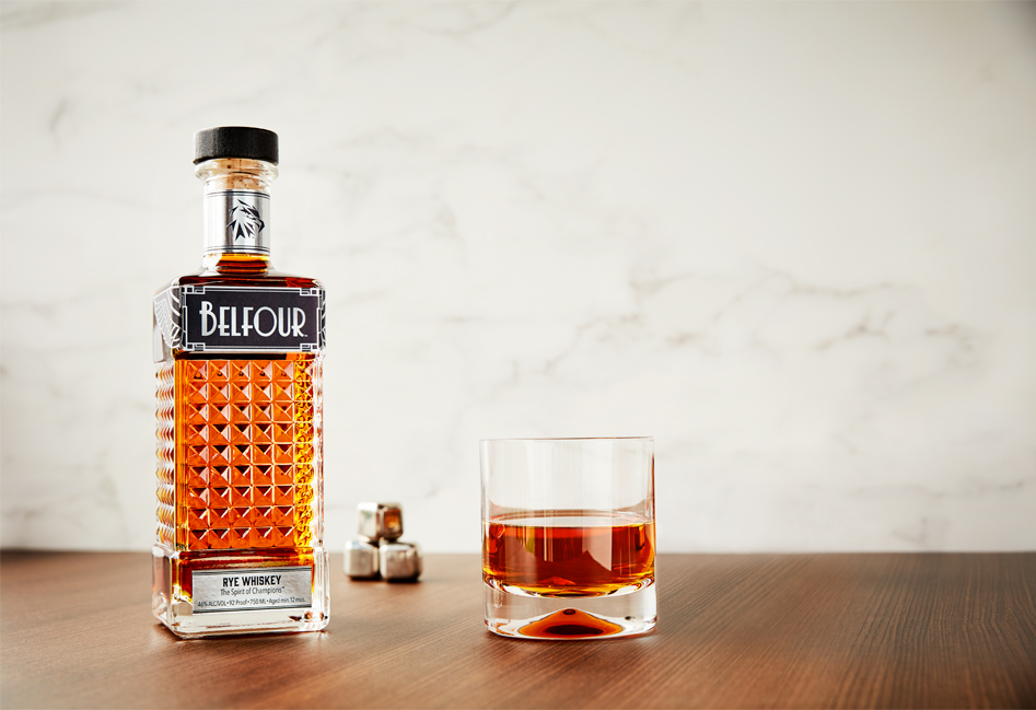Belfour Rye Whiskey bottle with metallic silver label with metal whiskey cubes and a low ball glass on a polished wood background with a marble background