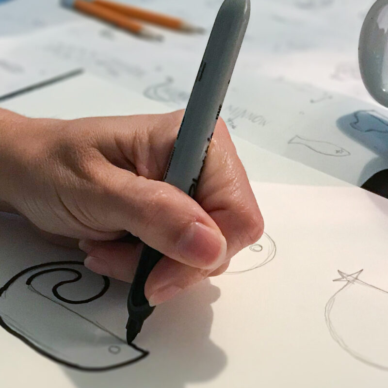 Graphic design sketching a logo with a black marker, pencils, sketches and an HCK2 coffee mug in the background